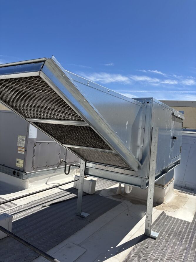 A large industrial HVAC unit with metal ductwork mounted on a rooftop under a clear blue sky.