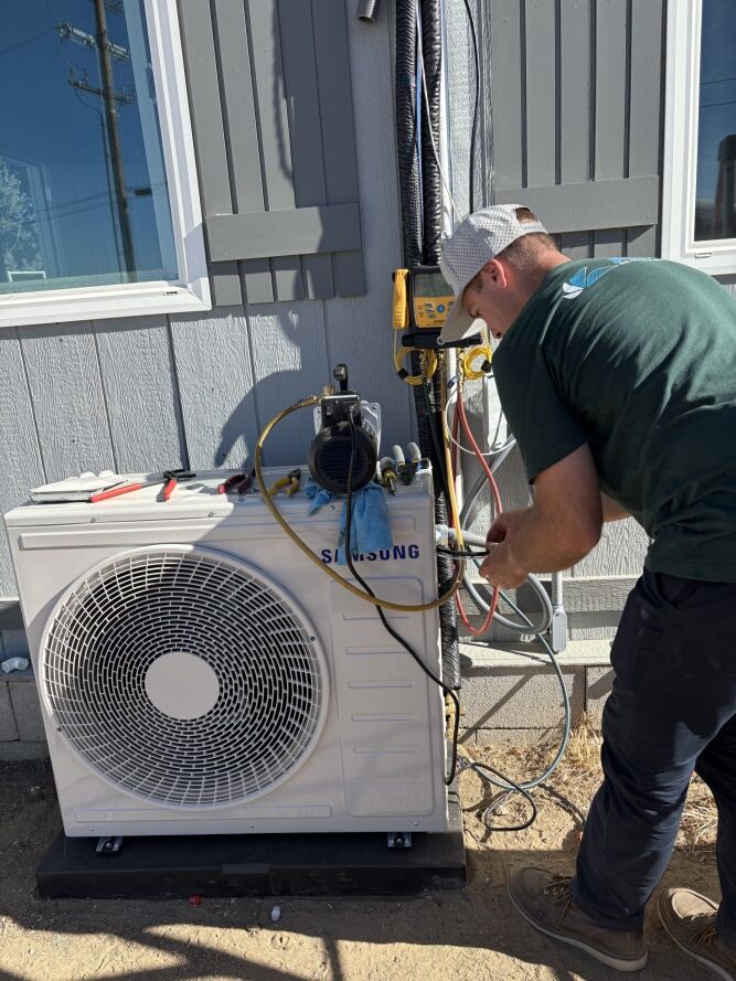 A technician in a green shirt services a Samsung outdoor HVAC unit, using tools and equipment attached to the system beside a gray building.