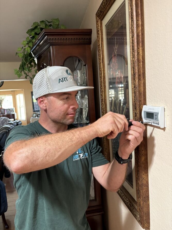 A man in a cap uses a screwdriver to adjust or install a thermostat on a wall in a residential setting.