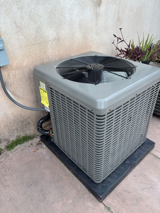 A gray outdoor air conditioning unit sits on a black pad next to a stucco wall, with plants and electrical wiring visible nearby.