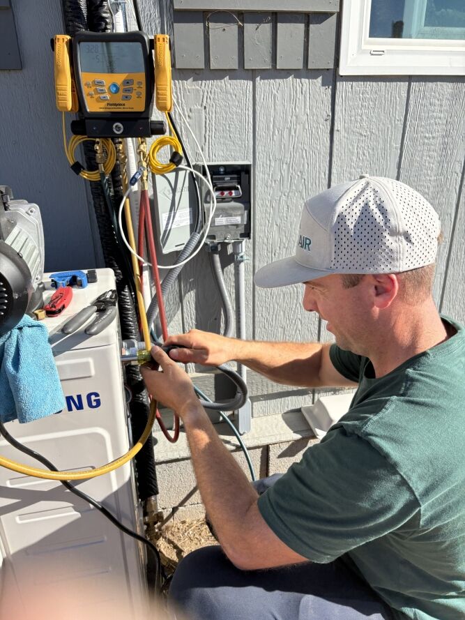 A technician in a cap uses tools to service an outdoor HVAC unit connected to gauges and wires, working next to a gray building.