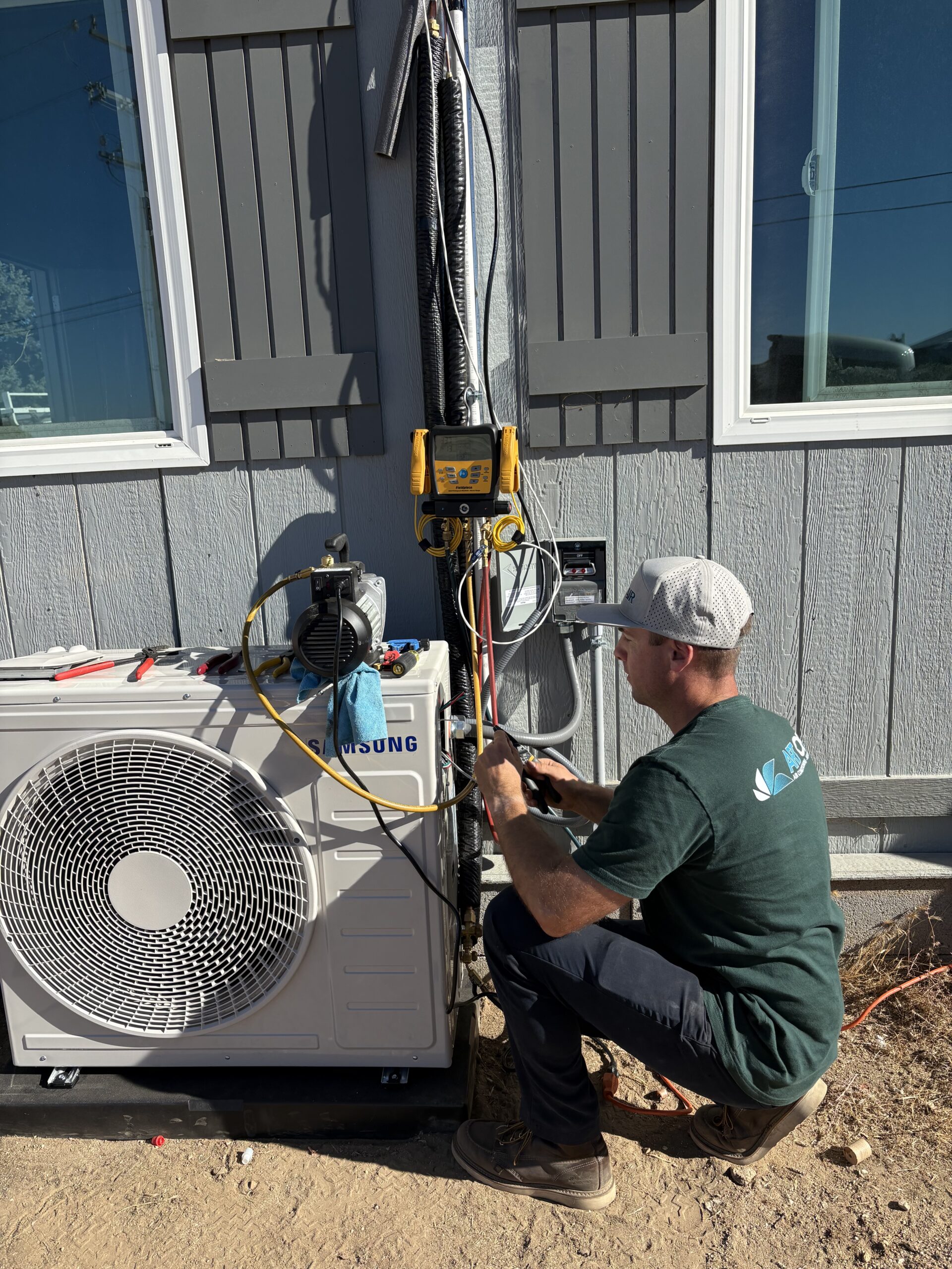 A technician kneels beside a building, servicing an outdoor air conditioning unit with various tools and gauges attached.