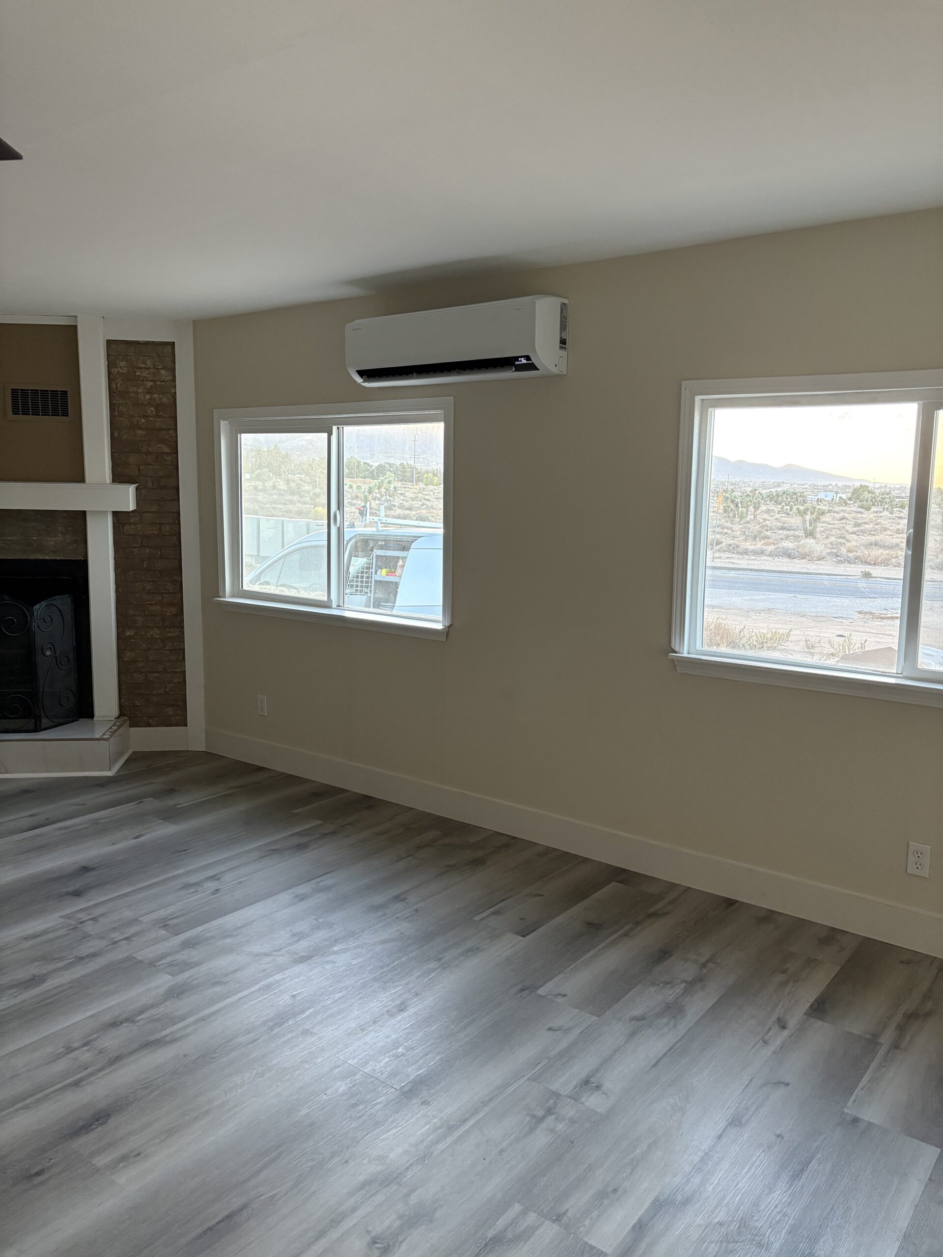 Empty room with light gray wood flooring, beige walls, two windows showing an outdoor view, a wall-mounted air conditioner, and a partial view of a fireplace.