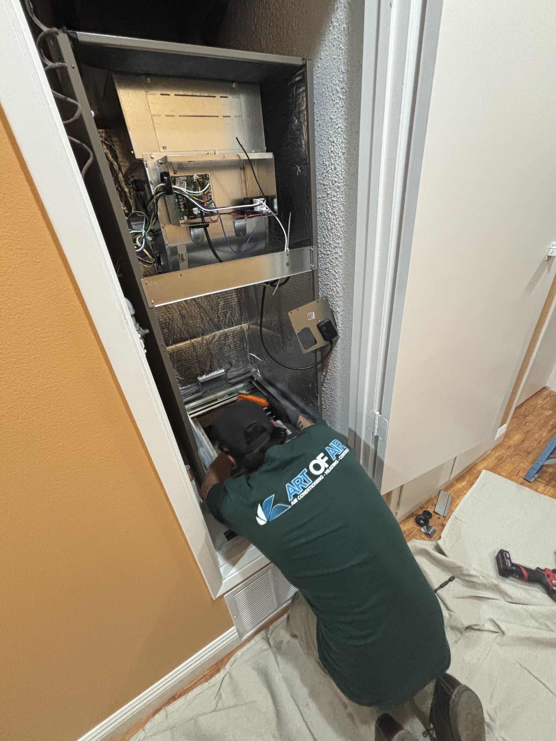 Technician kneeling on a drop cloth while working on wiring inside an open electrical panel mounted in a wall.