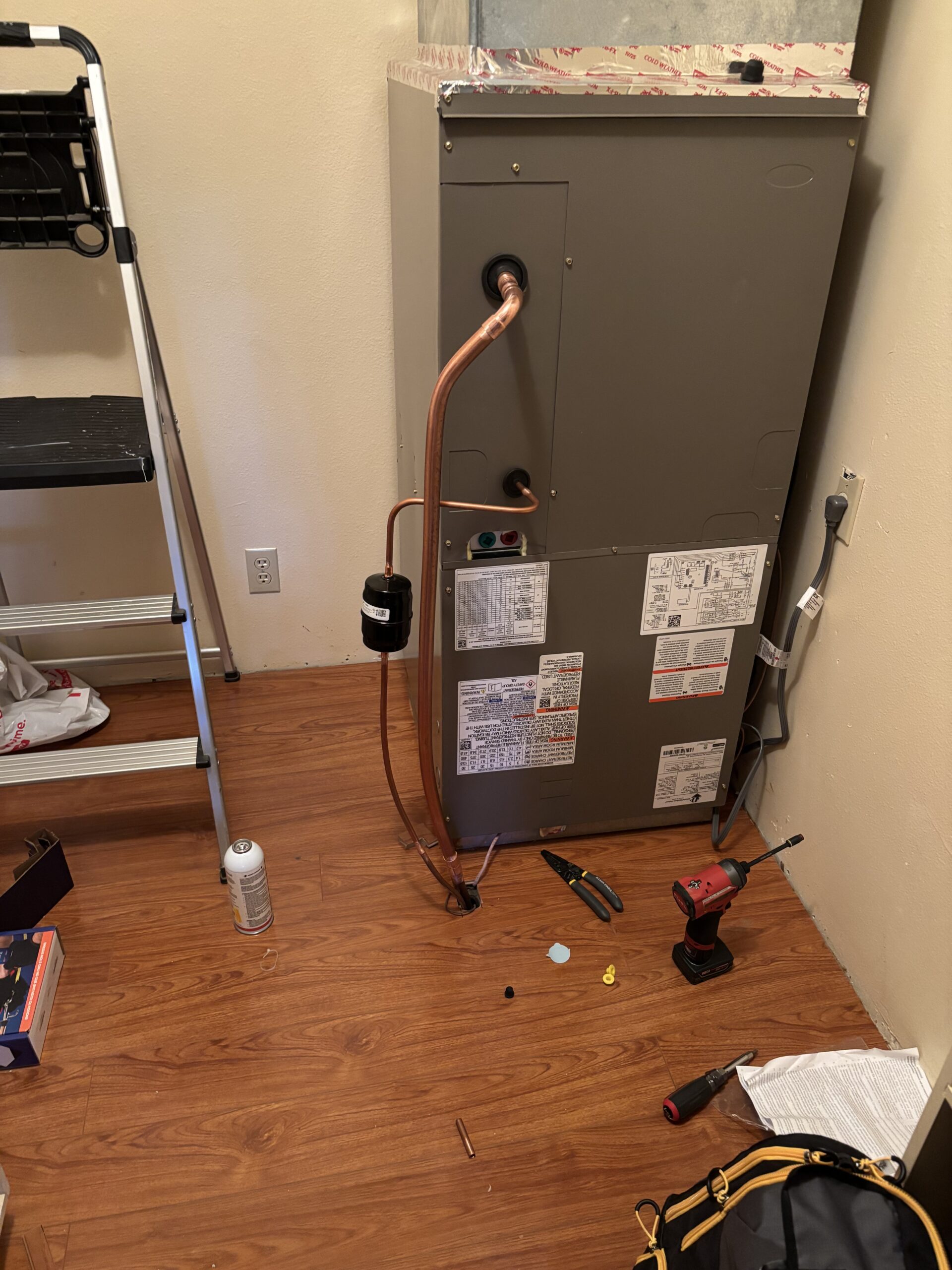 An HVAC unit with copper pipes installed, surrounded by tools, a drill, an open manual, and a step ladder in a room with wood flooring.