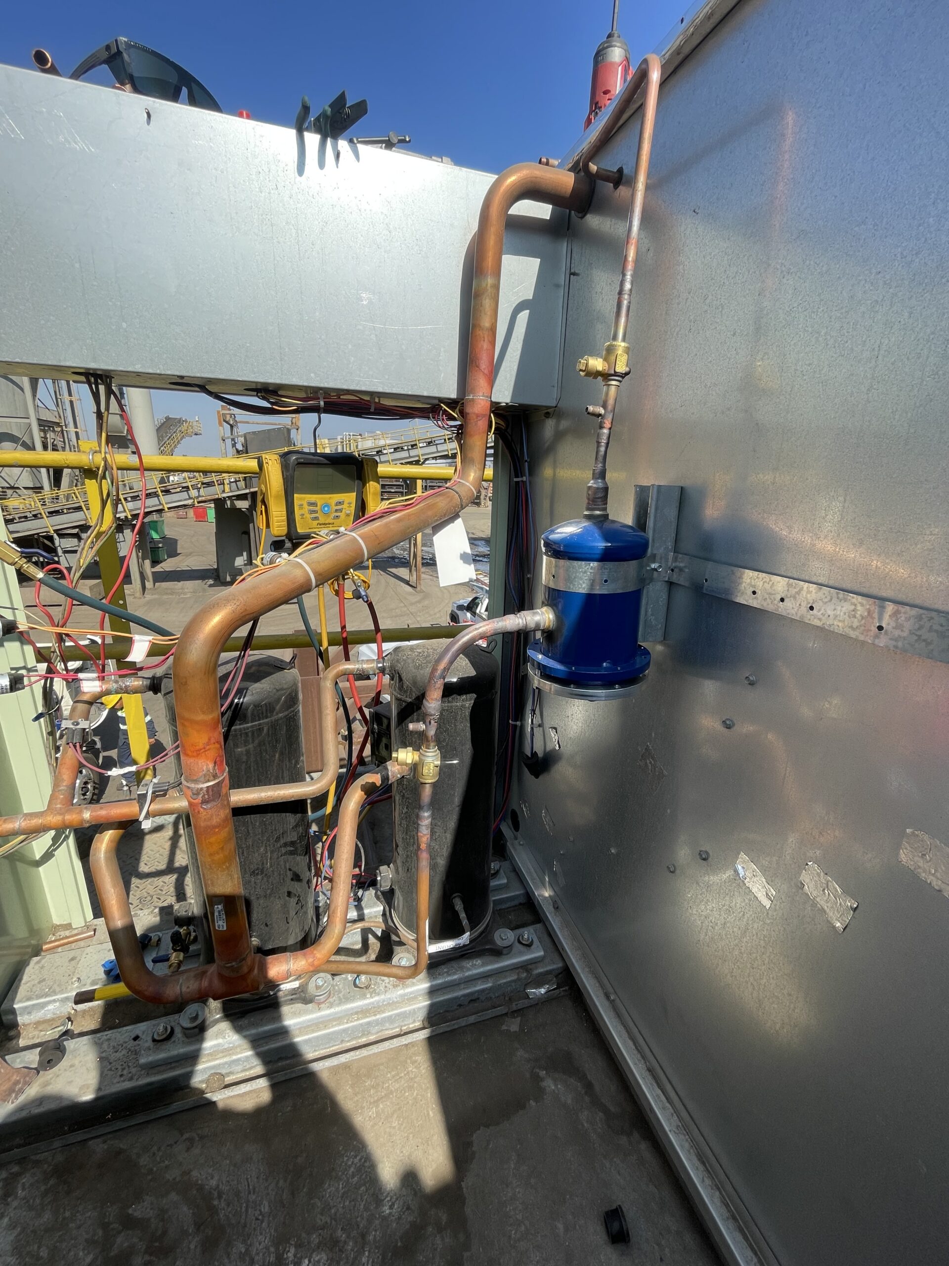 Copper pipes connected to HVAC equipment mounted on a metal panel, with a blue cylindrical filter-drier and electrical cables visible, outdoors under clear sky.