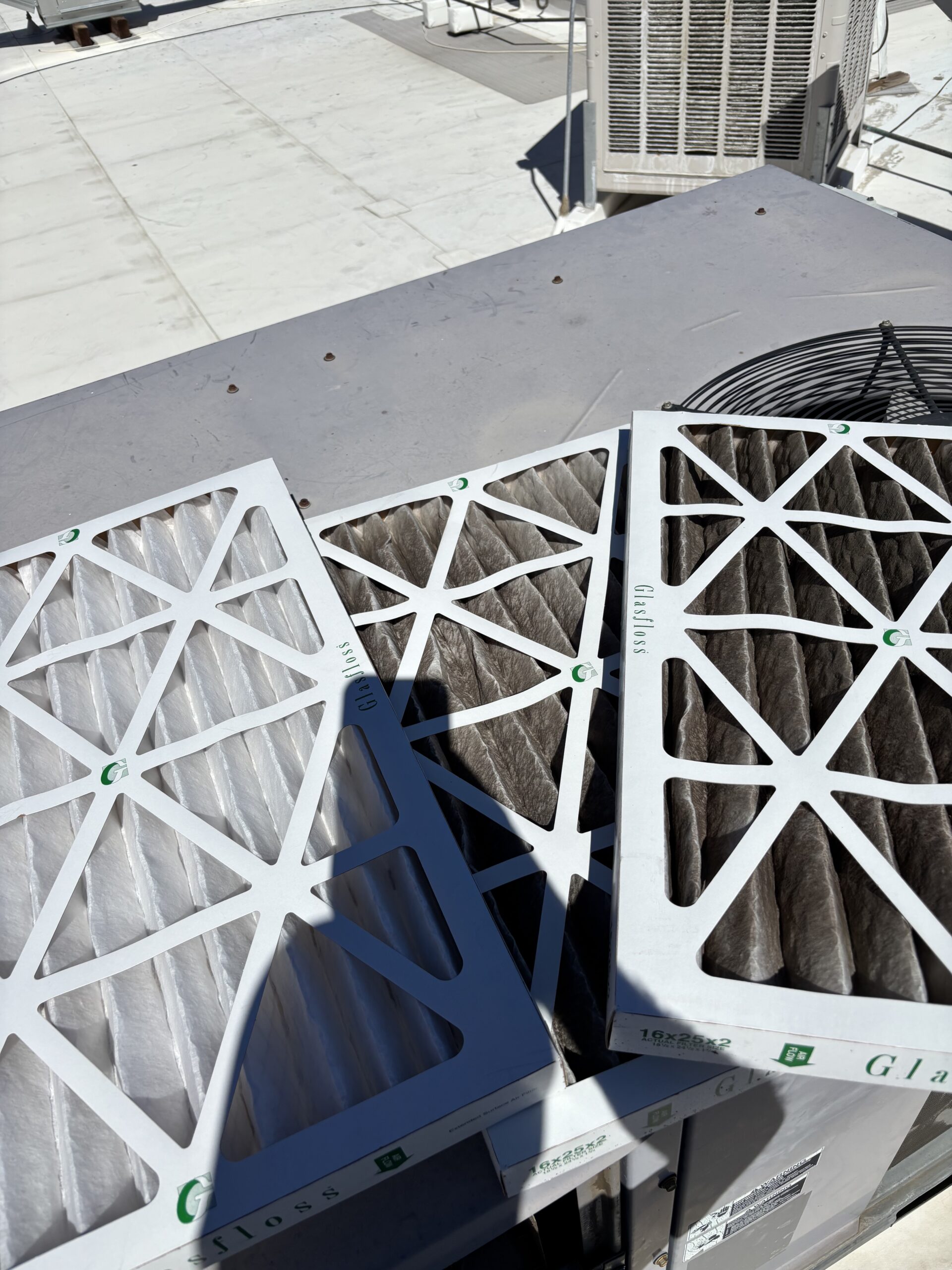 Three air filters, two dirty and one clean, are placed on top of a rooftop HVAC unit under direct sunlight.