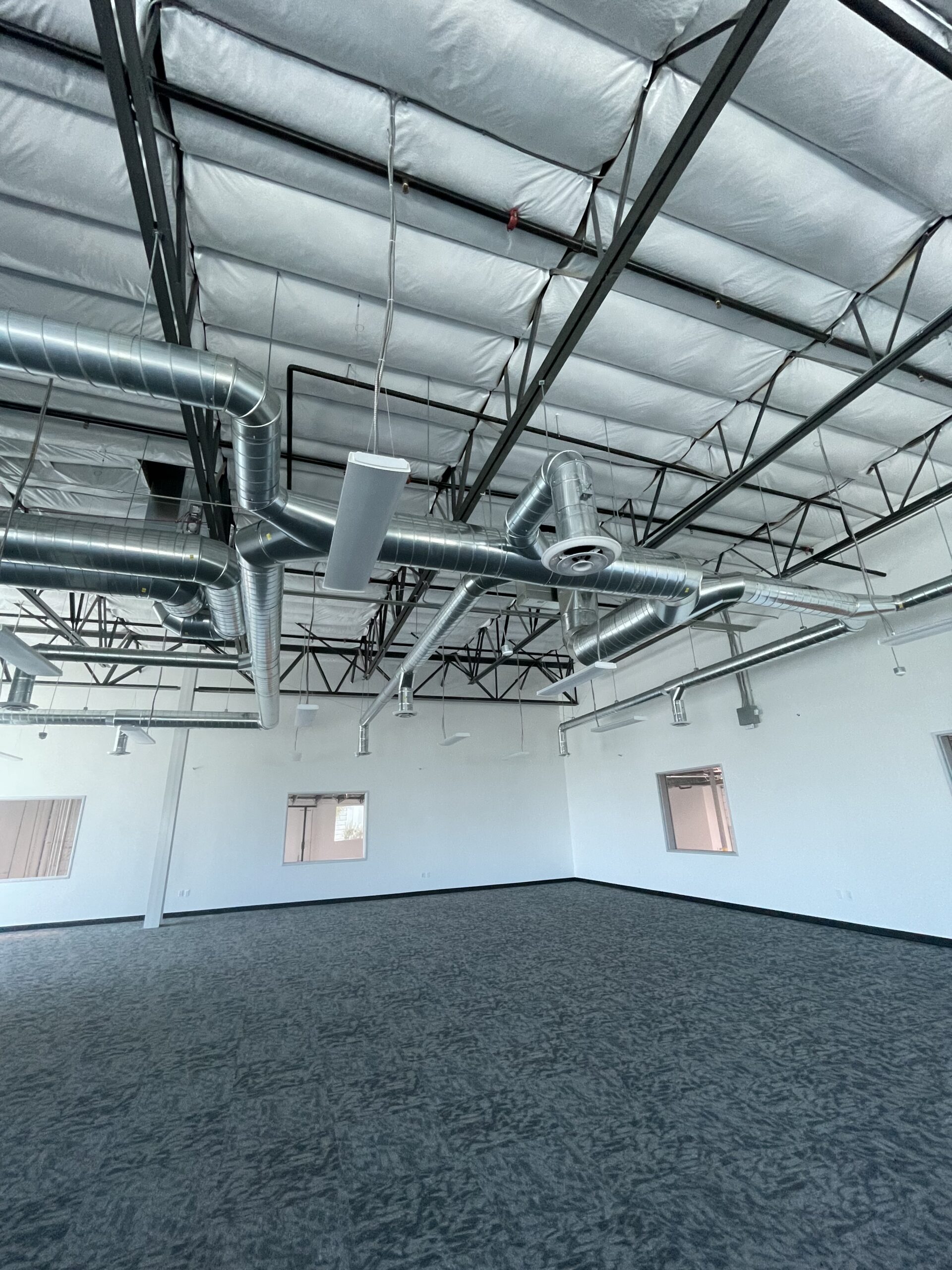 Large empty industrial room with gray carpet, white walls, windows, and exposed ceiling ducts and pipes.